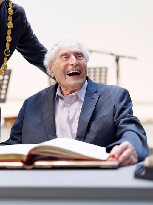 Ein sehr alter Mann sitzt lachend an einem großen Tisch. 