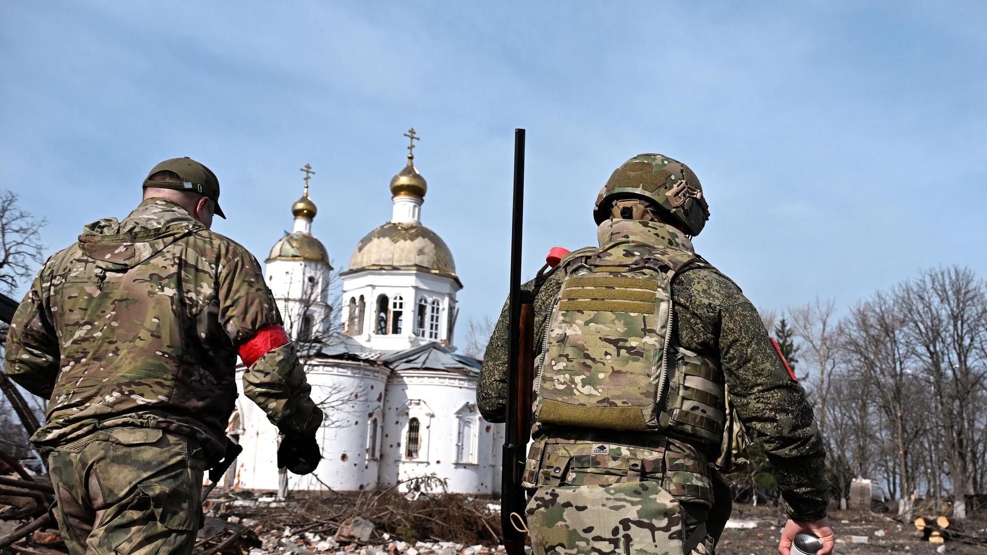 Zwei russische Soldaten von hinten vor einer beschädigten, weißen orthodoxen Kirche mir goldener Kuppel in der russischen Region Kursk. 