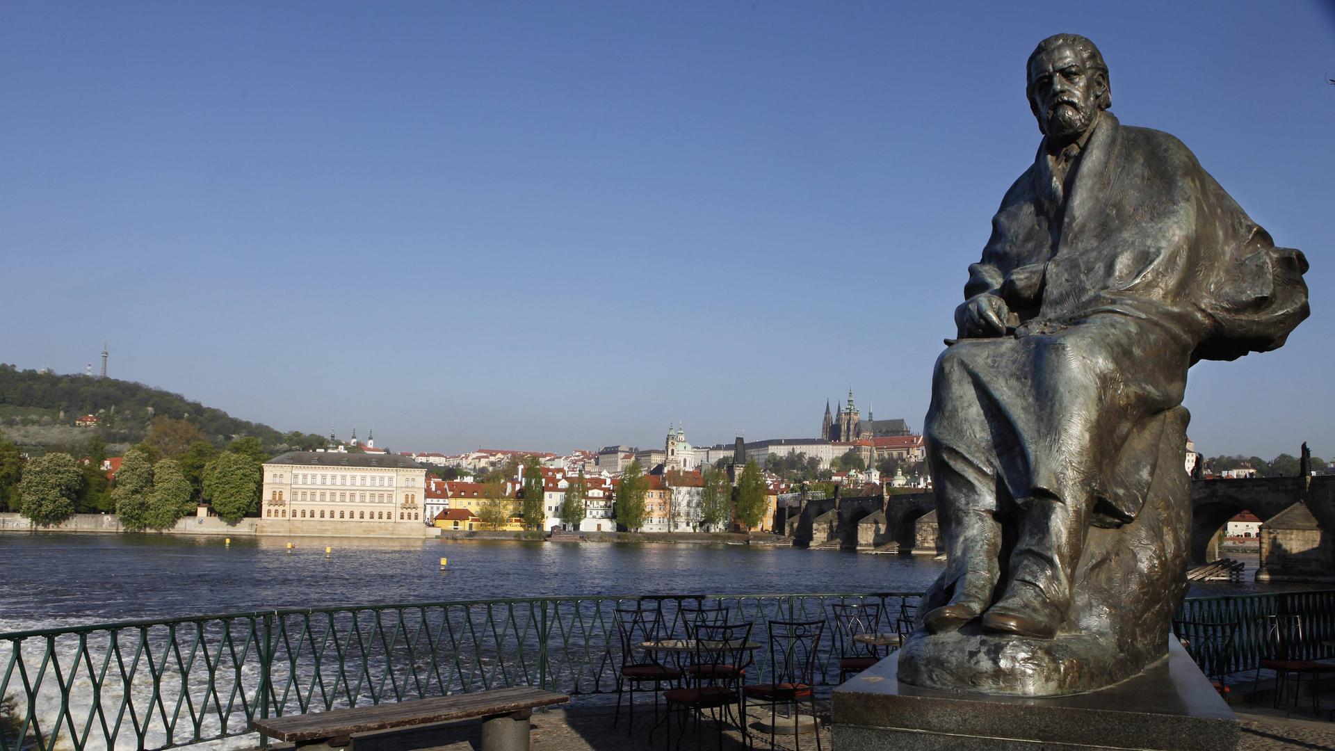 Blick auf Sockel und darauf sitzender Smetana-Figur mit wehendem Mantel, im Hindergrund die Karlsbrücke und der typische Altstadt-Blick von Prag. 