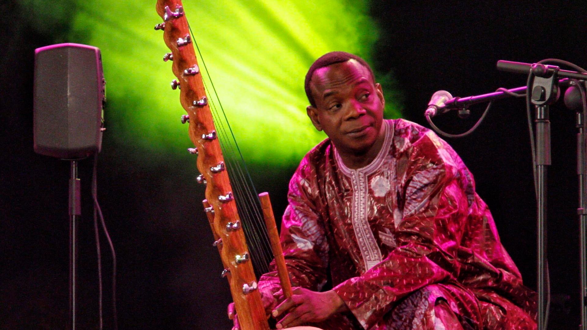 Musiker Toumani Diabate aus Mali bei einem Auftritt in Spanien. Er sitzt in einem bunten Gewand, rechts ist es Mikroständer zu sehen, und hält sein Instrument.