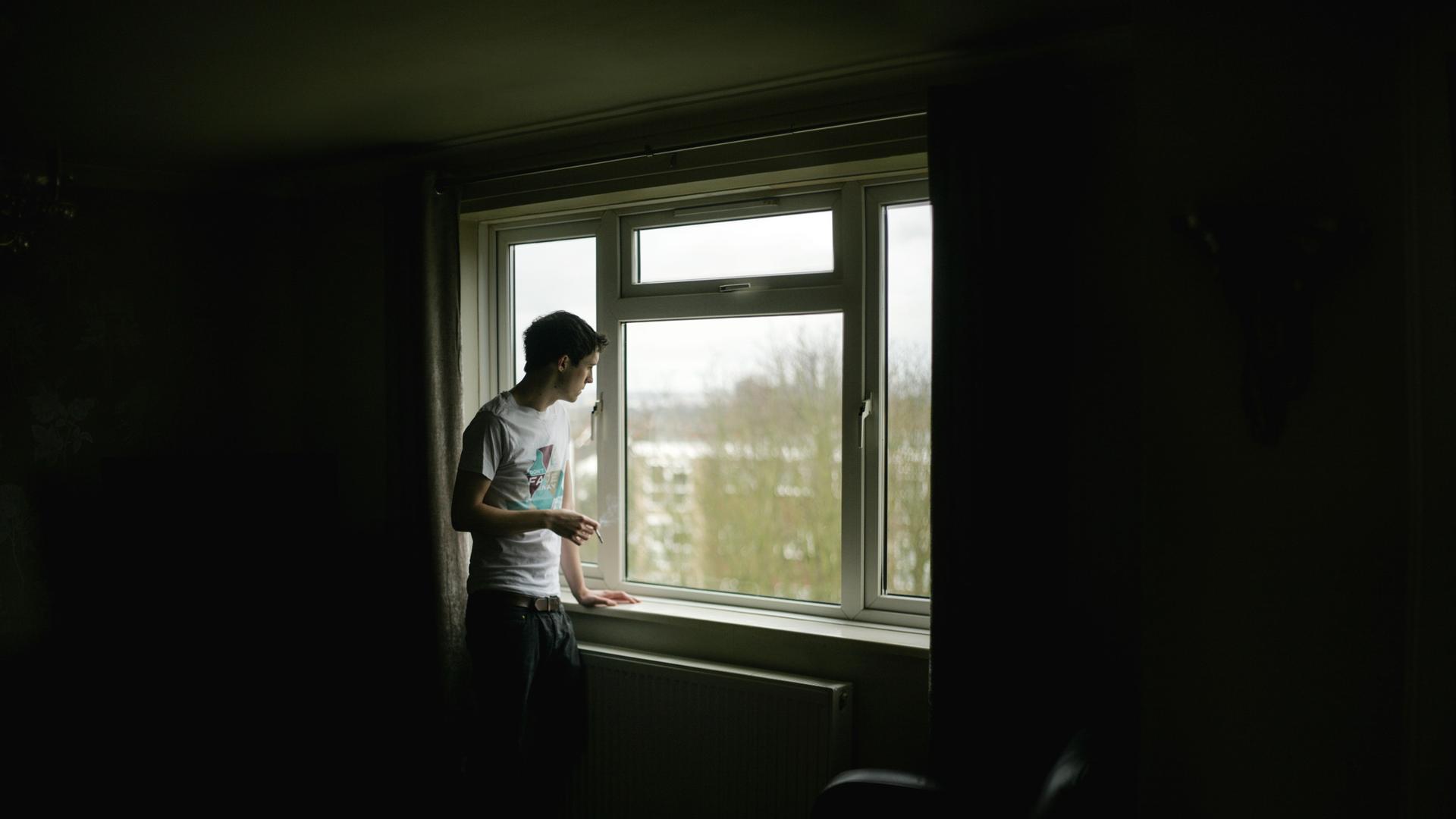 Ein junger Mann steht alleine am Fenster und raucht.