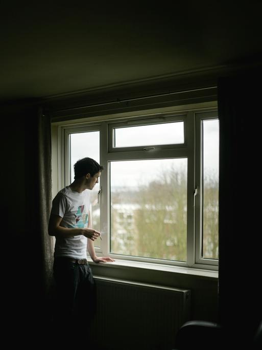 Ein junger Mann steht alleine am Fenster und raucht.