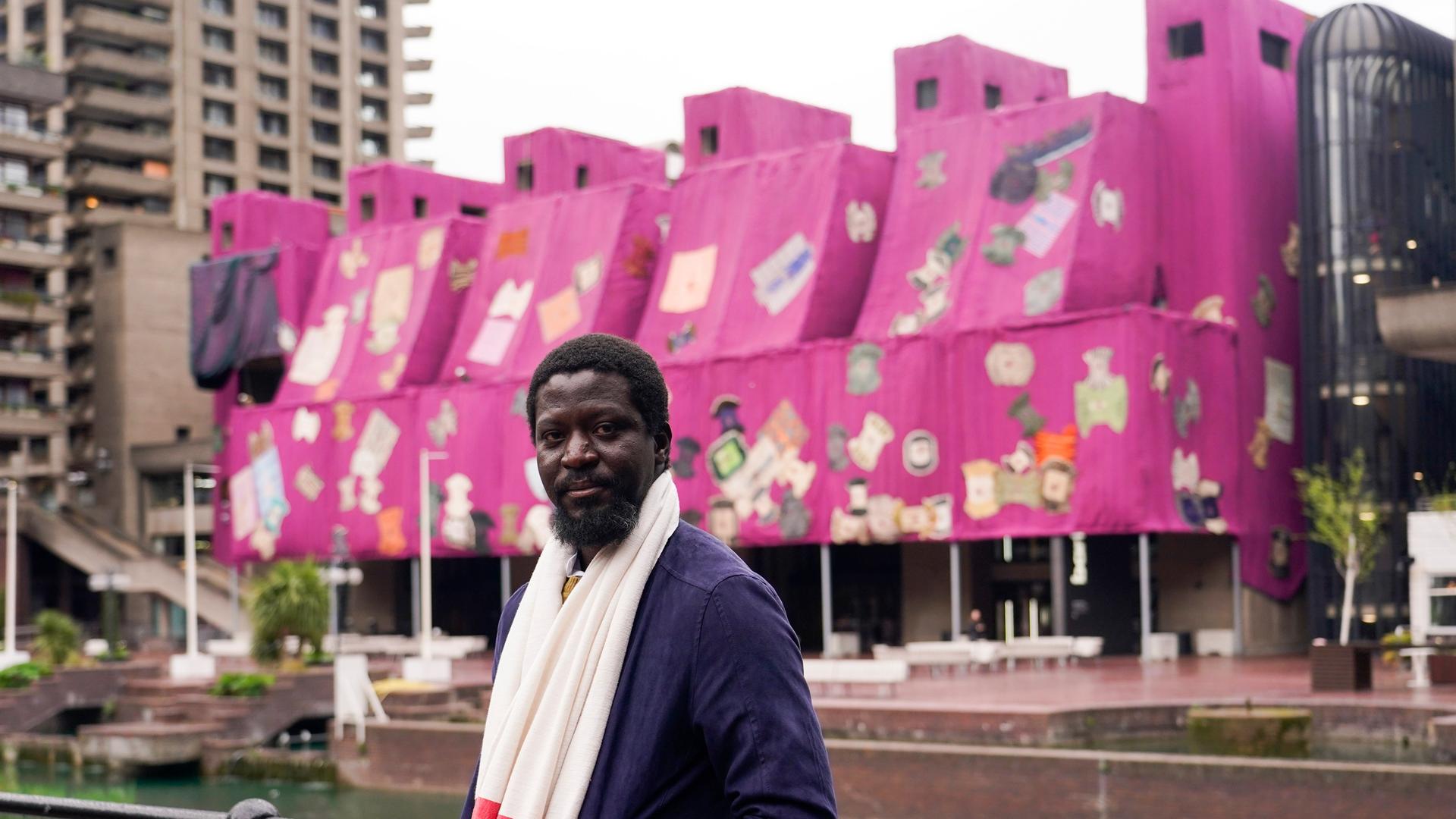 Ibrahim Mahama steht vor einem pink und lila umhüllten Betongebäude. poses for photographs in front of his artwork Purple Hibiscus, outside the Barbican Centre, in London, Tuesday, April 9, 2024. Envelopin Ibrahim Mahama steht vor einem pink und lila umhüllten Betongebäude. poses for photographs in front of his artwork Purple Hibiscus, outside the Barbican Centre, in London, Tuesday, April 9, 2024. Envelopin