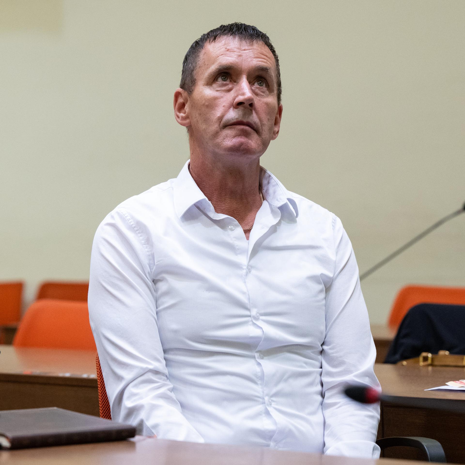 Genditzki im weißen Hemd blickt mit ernstem Blick nach vorne. Im Hintergrund unscharf Gerichtsbänke und Stühle. 