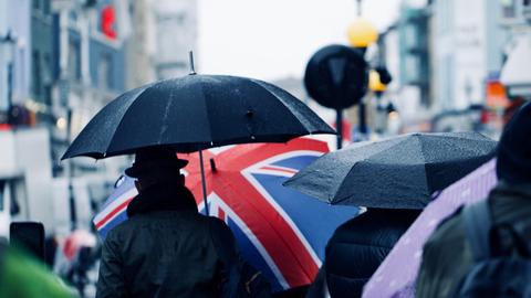 Straßenszene in London: Von hinten sieht man Menschen mit Schirmen, eine Person trägt einen Schirm, der mit der britischen Flagge bedruckt ist.