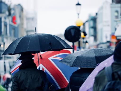 Straßenszene in London: Von hinten sieht man Menschen mit Schirmen, eine Person trägt einen Schirm, der mit der britischen Flagge bedruckt ist.