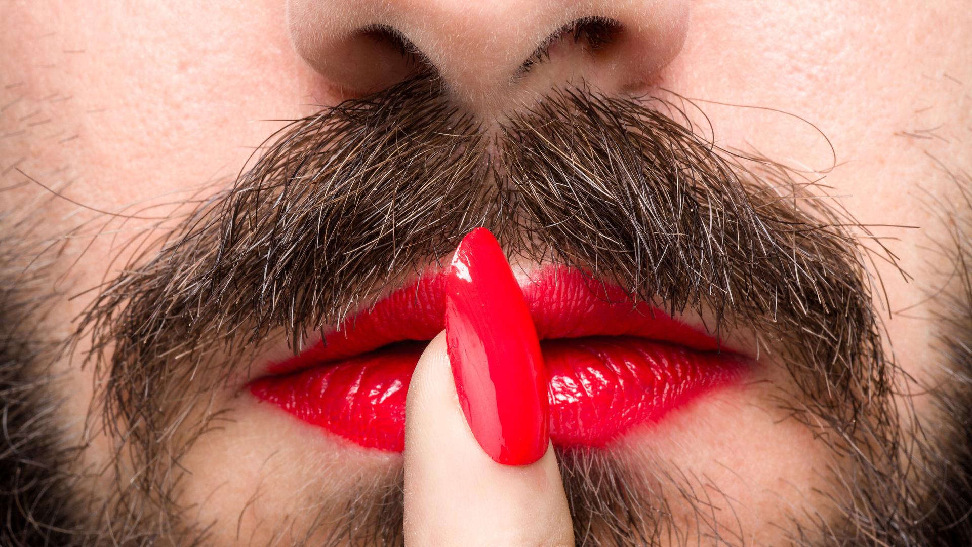 Detailansicht eines Mannes mit Bart, rotem Lippenstift und lackiertem Fingernagel. Detailansicht eines Mannes mit Bart, rotem Lippenstift und lackiertem Fingernagel.