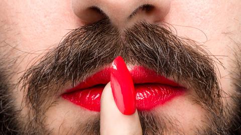 Detailansicht eines Mannes mit Bart, rotem Lippenstift und lackiertem Fingernagel.