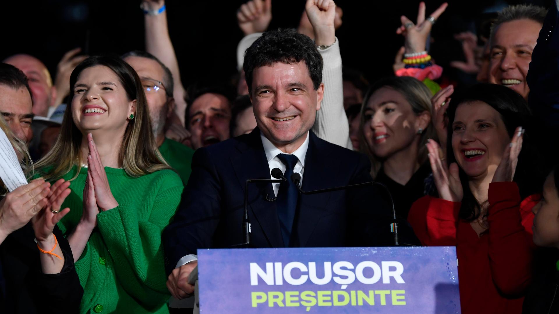 Nicusor Dan steht an einem Redner-Pult und lacht. Auf dem Pult steht "Nicusor Präsident" auf rumänisch. Neben und hinter ihm stehen jubelnde Menschen. Nicusor Dan steht an einem Redner-Pult und lacht. Auf dem Pult steht "Nicusor Präsident" auf rumänisch. Neben und hinter ihm stehen jubelnde Menschen.