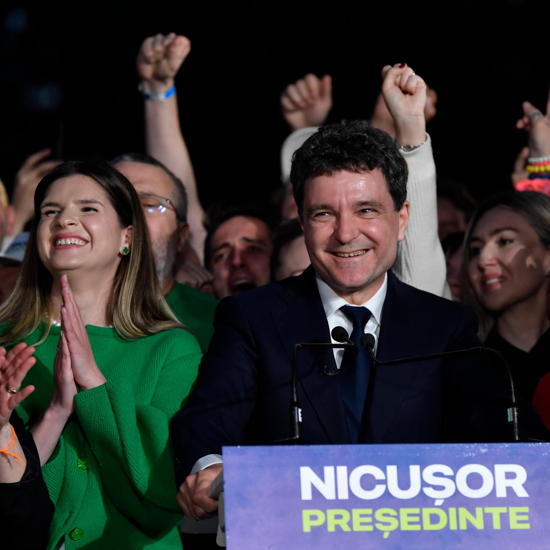 Nicusor Dan steht an einem Redner-Pult und lacht. Auf dem Pult steht "Nicusor Präsident" auf rumänisch. Neben und hinter ihm stehen jubelnde Menschen.