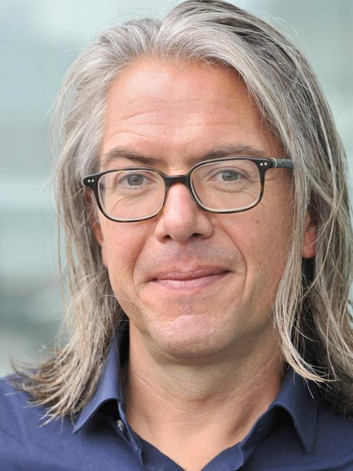 Ein rund fünfzigjähriger Mann mit schulterlange, grauen Haaren, Steffen Kopetzky schaut in die Kamera. Er trägt ein blaues Hemd und eine Brille mit dunklem Rand.