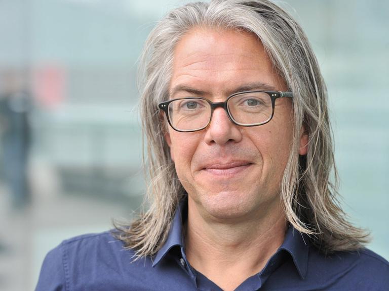 Ein rund fünfzigjähriger Mann mit schulterlange, grauen Haaren, Steffen Kopetzky schaut in die Kamera. Er trägt ein blaues Hemd und eine Brille mit dunklem Rand.