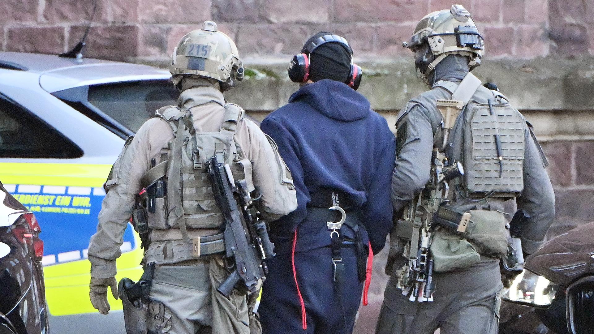 Der Mann im blauen Overall und mit Ohrenschutz wird von zwei vermummten Polizisten in Kampfmonitur abgeführt. Alle sind von hinten fotografiert. Im Hintergrund eine Wand des Bundesgerichtshofs und ein Polizeiauto. 