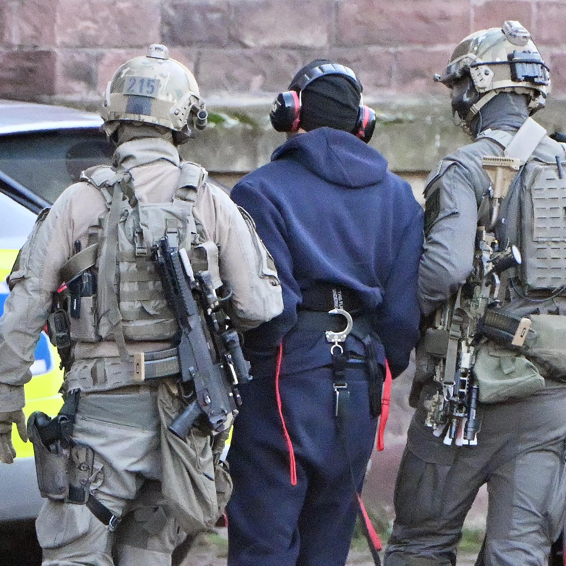 Eine Person wird von zwei vermummten Personen abgeführt. Alle sind von hinten fotografiert. 