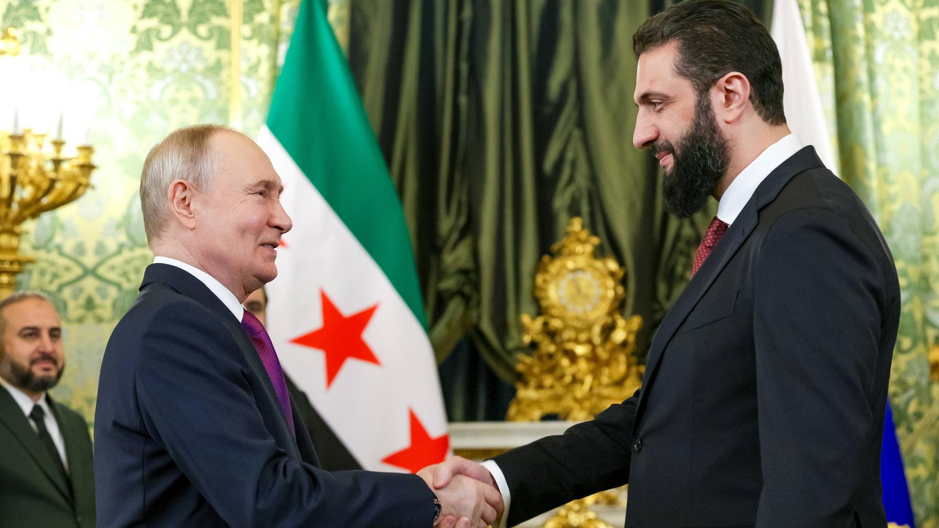 Die beiden begrüßen sich lächelnd mit Handschlag. Dahinter eine Wand mit einem grünen Vorhang und einer syrischen Flagge. Die beiden begrüßen sich lächelnd mit Handschlag. Dahinter eine Wand mit einem grünen Vorhang und einer syrischen Flagge.