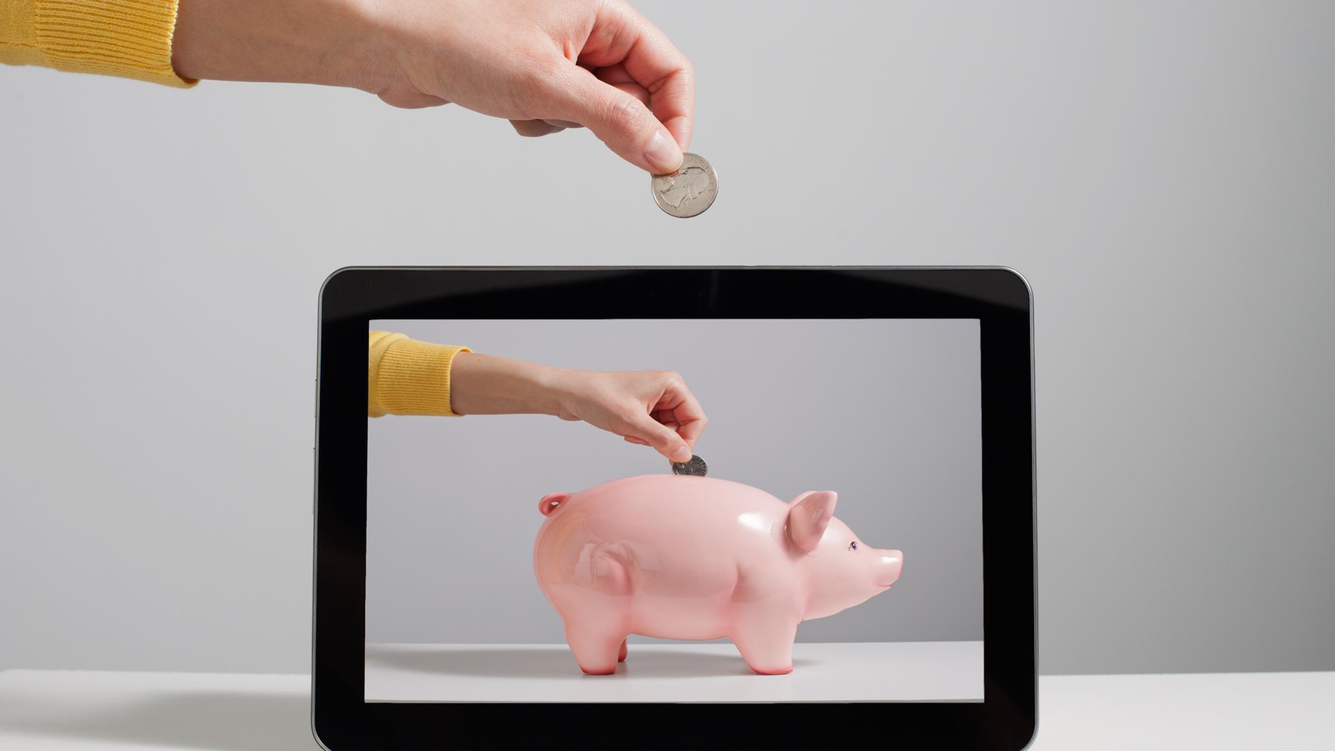 Eine Hand wirft Geld in ein Sparschwein, welches nur noch auf dem Bildschirm auftauscht.