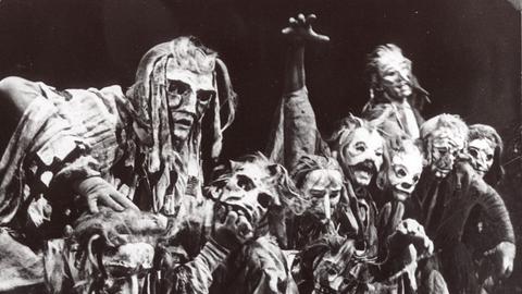 Mehrere markant-verzerrte Masken drängen sich auf dem historischen Foto in schwarz-weiß in einer Reihe eng nebeneinander. 