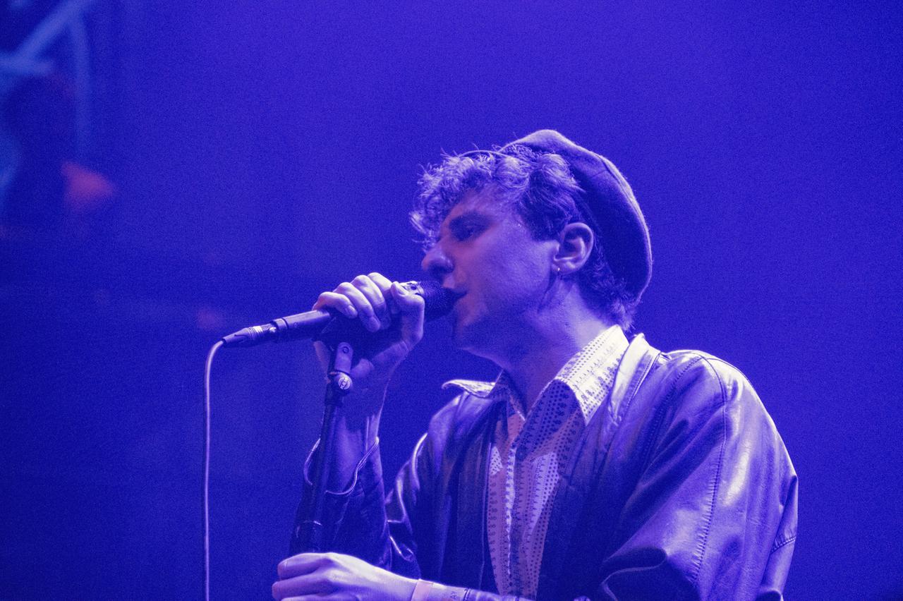 Tobias Bamborschke singt auf einer lilafarbenen Bühne mit Mütze auf dem Kopf, während er sein Mikrofon festhält.