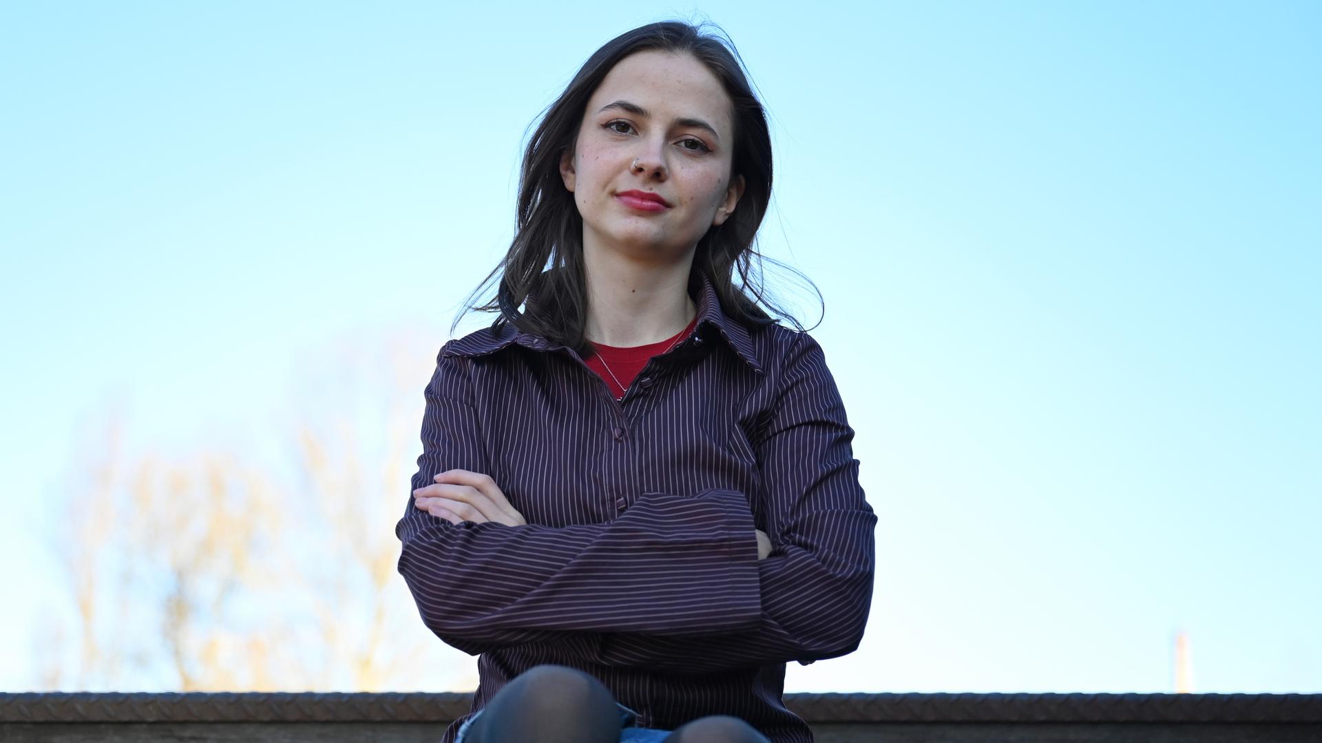 Theresia Crone sitzt im Park am Gleisdreieck in Berlin. Vor blauem Himmel im Hintergrund hält sie die Arme verschränkt und blickt direkt in die Kamera.