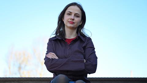 Theresia Crone sitzt im Park am Gleisdreieck in Berlin. Vor blauem Himmel im Hintergrund hält sie die Arme verschränkt und blickt direkt in die Kamera.