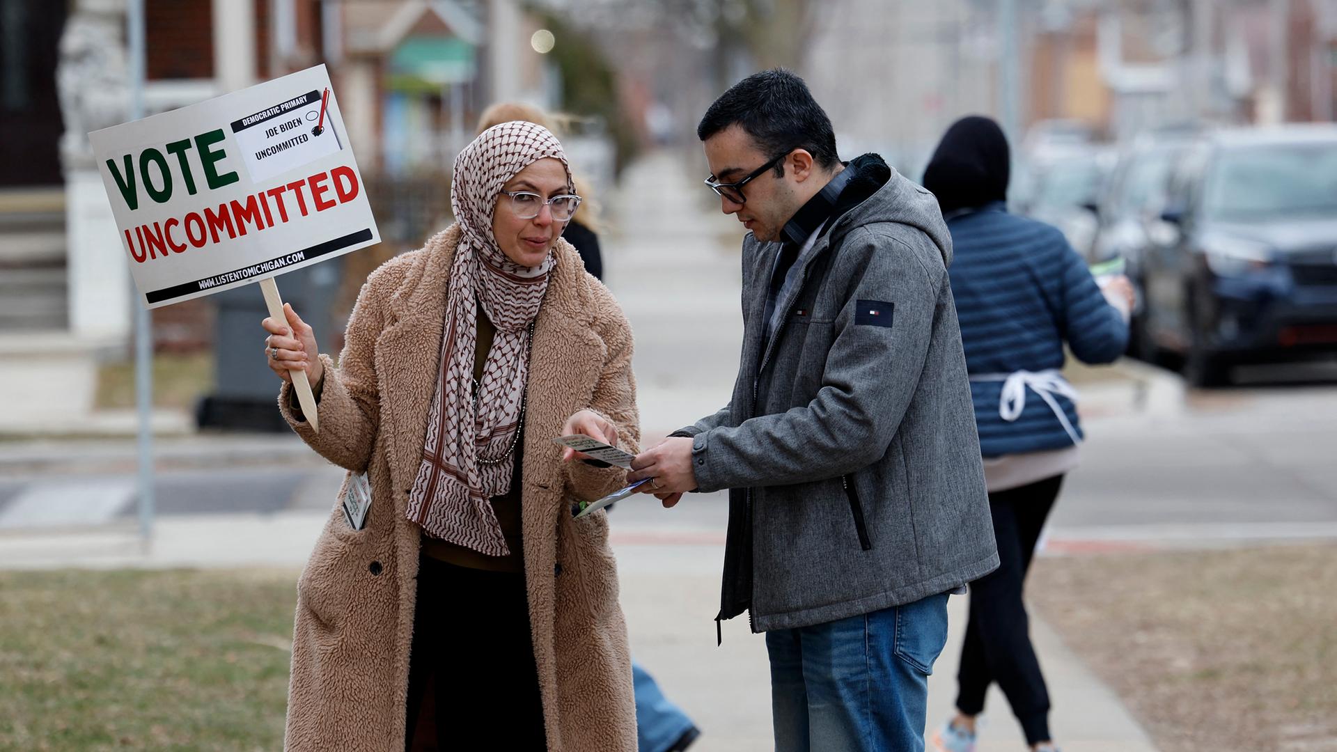 Eine muslimische Frau spricht auf der Straße mit einem Mann. Sie hält ein Plakat in der Hand mit der Aufschrift "Vote uncommitted" (deutsch: "Wähle neutral").