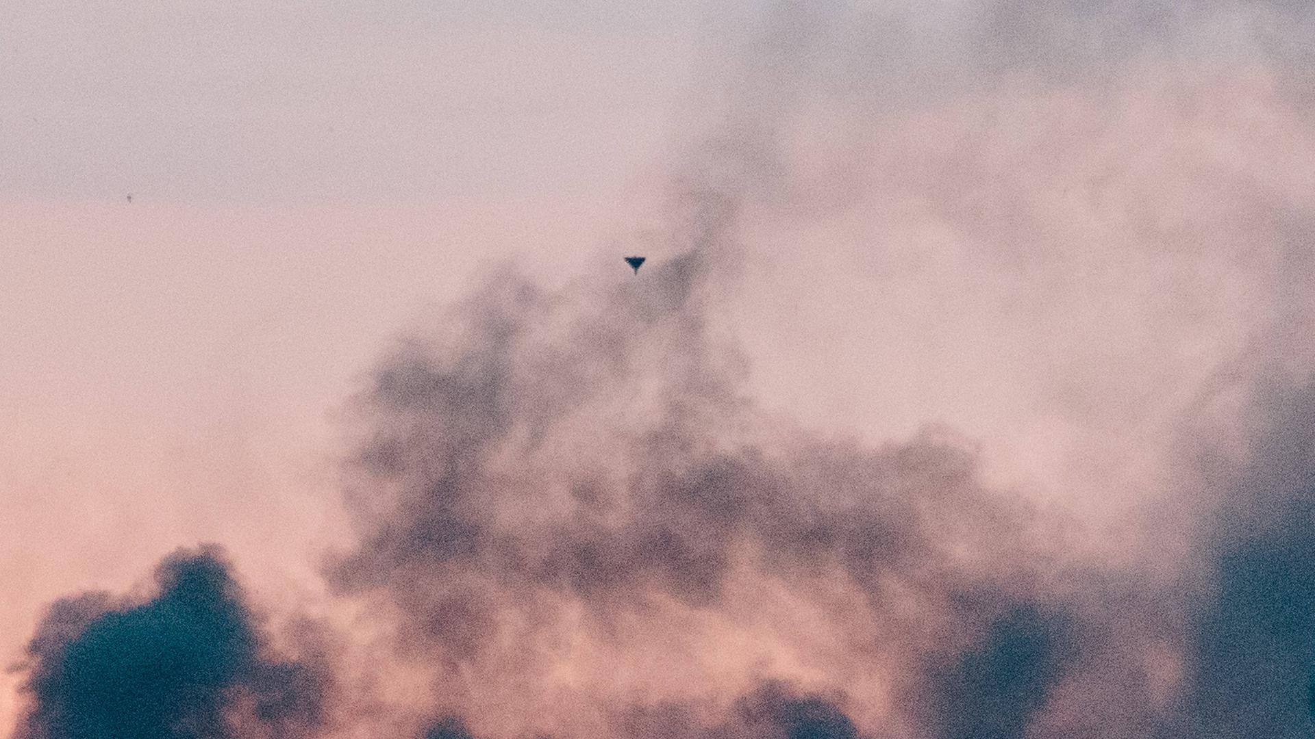 Eine von Russland eingesetzte Shahed-Drohne ist klein am rosafarbenen Himmel über Charkiw zu sehen. Im Vordergrund sind dunkle Rauchschwaden. Eine von Russland eingesetzte Shahed-Drohne ist klein am rosafarbenen Himmel über Charkiw zu sehen. Im Vordergrund sind dunkle Rauchschwaden.