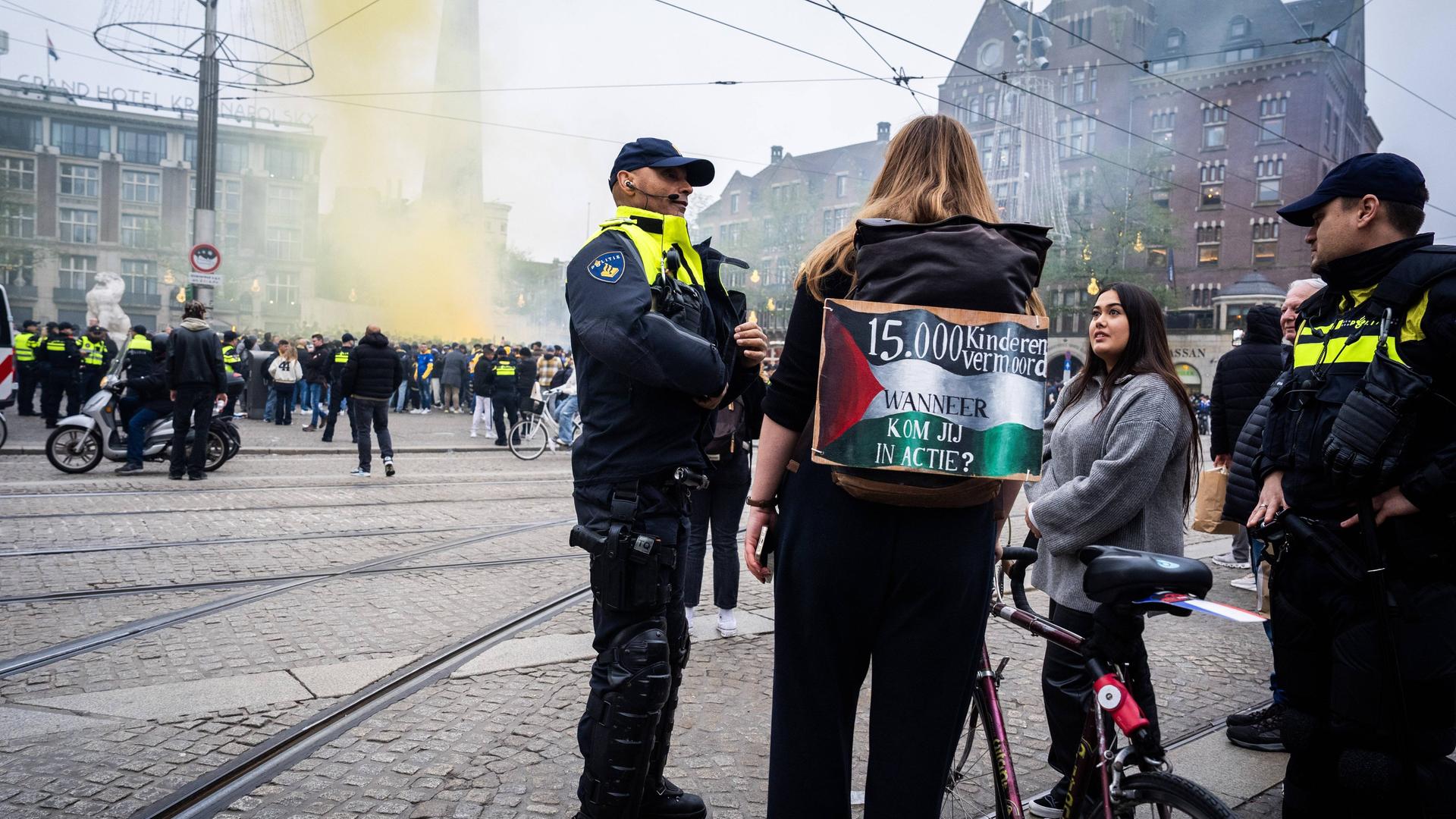 Polizisten und ein pro-palästinensischer Demonstrant sprechen in Amsterdam miteinander, im Hintergrund sind Fans des israelischen Fußballklubs Makkabi Tel Aviv. Gelbe Rauchschwaden steigen dort hoch. 