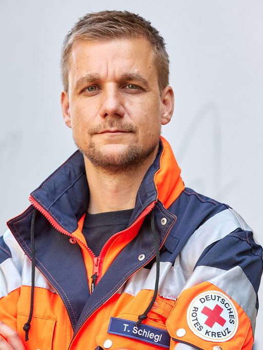 Ein Mann Mitte 40 mit kurzen Haaren und Kinnbart, Tobias Schlegl, steht in der Jacke eines Notfallsanitäters vor einer weißen Wand und blickt in die Kamera.