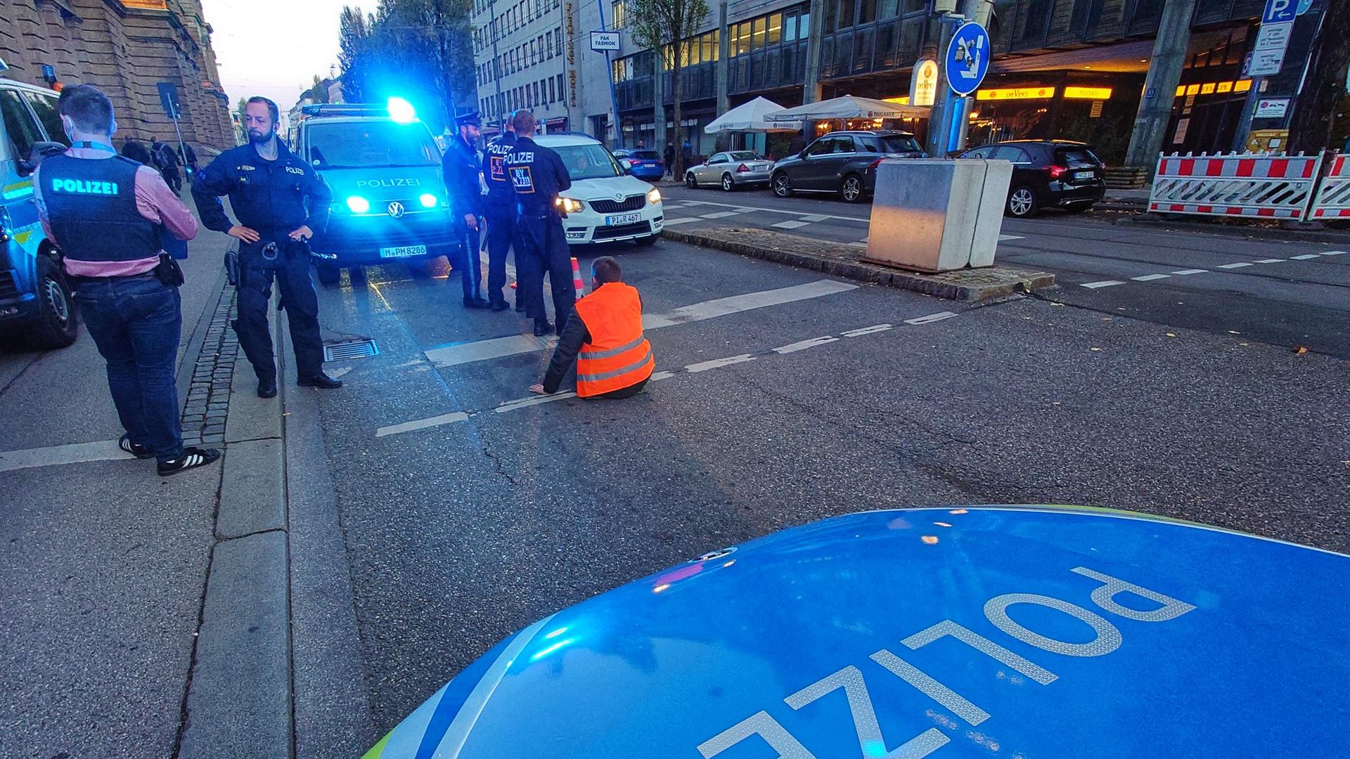 Ein Klimaaktivist hat sich am 7. November 2022 auf einer Straße in München an den Asphalt geklebt. Er träge eine rote Weste, die leuchtet. Um ihn herum stehen mit etwas Abstand mehrere Polizeiwagen und Polizisten, teilweise ist das Blaulicht an.