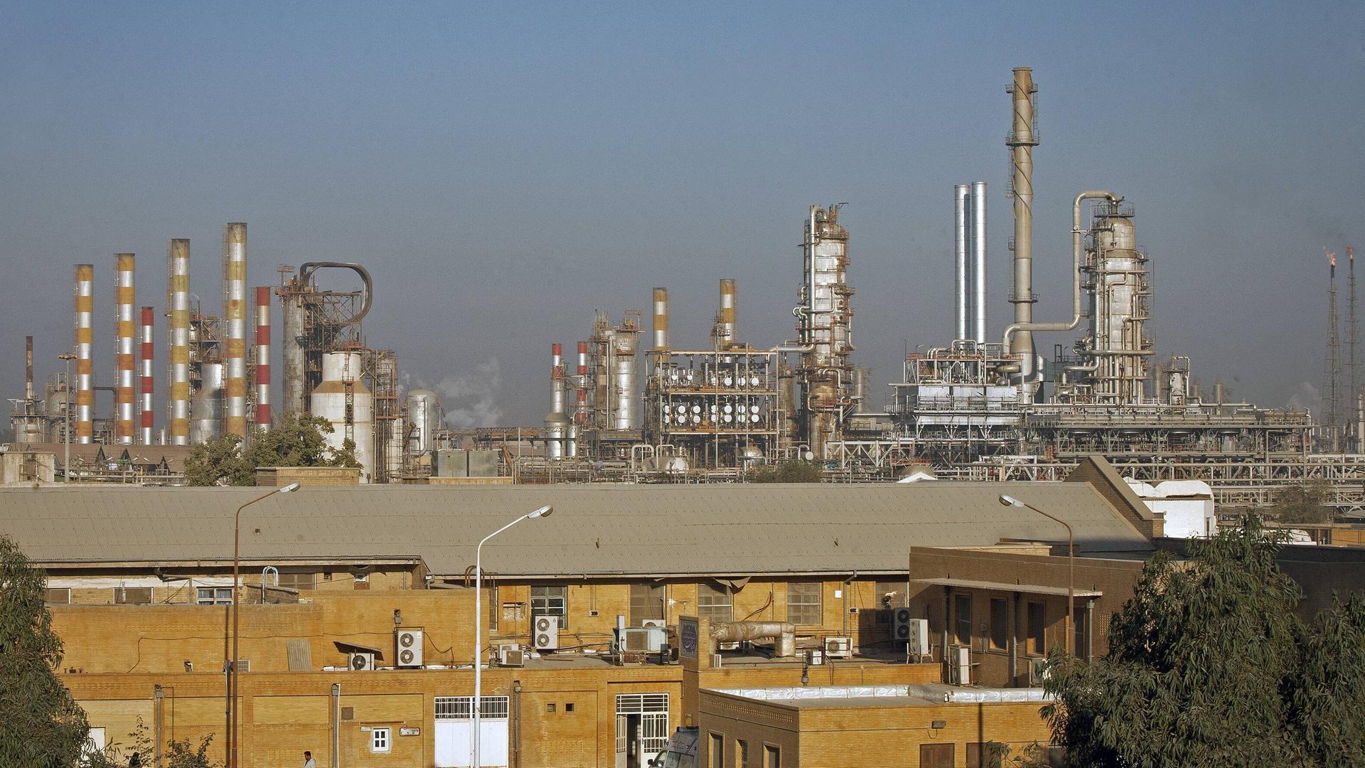Blick auf die Raffinerie aus größerer Entfernung. Mehrere Schlote ragen in den blauen Himmel. Im Vordergrund ein Gebäude aus hellbraunem Backstein. Blick auf die Raffinerie aus größerer Entfernung. Mehrere Schlote ragen in den blauen Himmel. Im Vordergrund ein Gebäude aus hellbraunem Backstein.