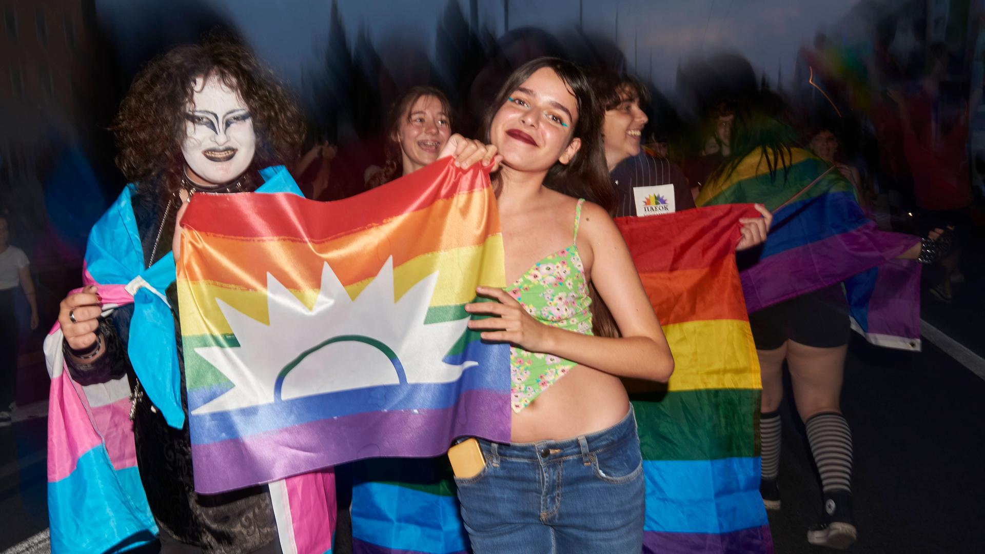 Zwei Frauen halten eine Regenbogenfahne. Das Bild ist in der Dunkelheit aufgenommen. Im Hintergrund sind weitere Menschen. Zwei Frauen halten eine Regenbogenfahne. Das Bild ist in der Dunkelheit aufgenommen. Im Hintergrund sind weitere Menschen.