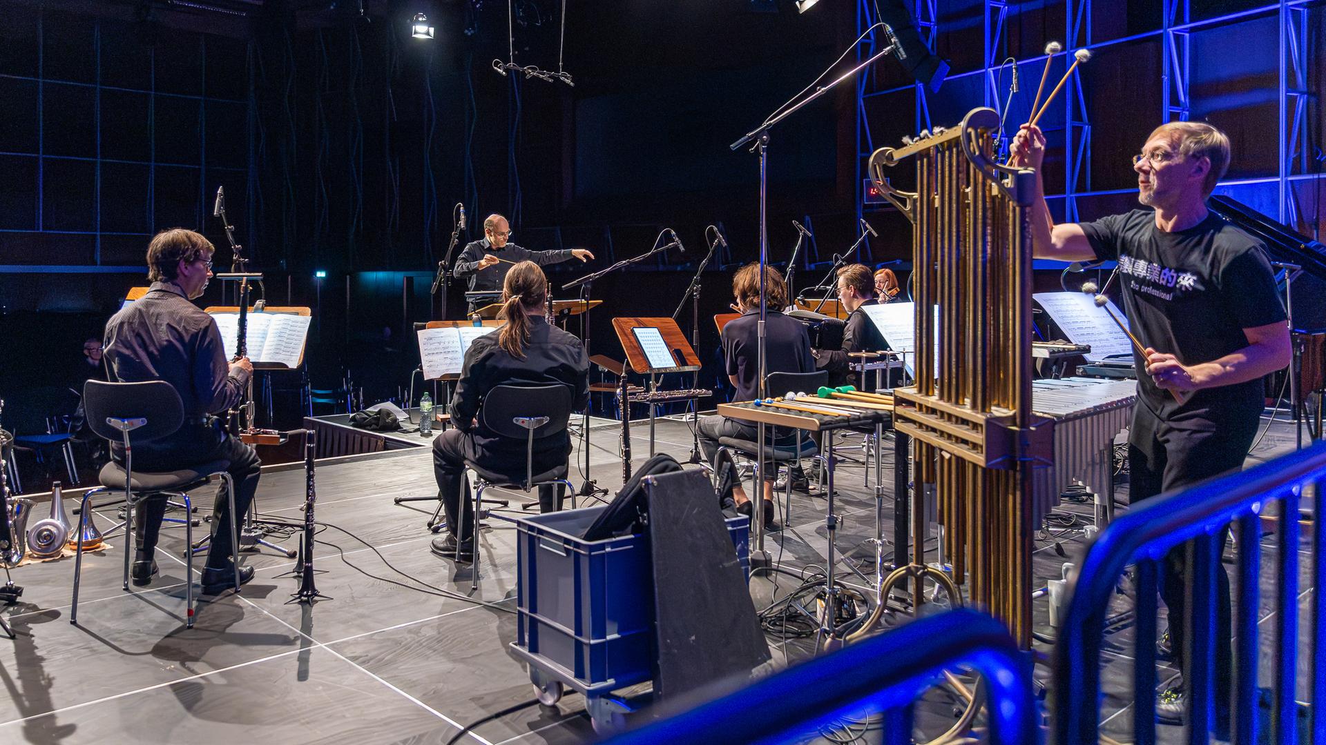 Die Musiker vom E-MEX Ensemble sind zum Teil von hinten, teils von der Seite zu sehen, wie sie gemeinsam spielen. Im Vordergrund stehen große Röhren des Percussions-Musikers.