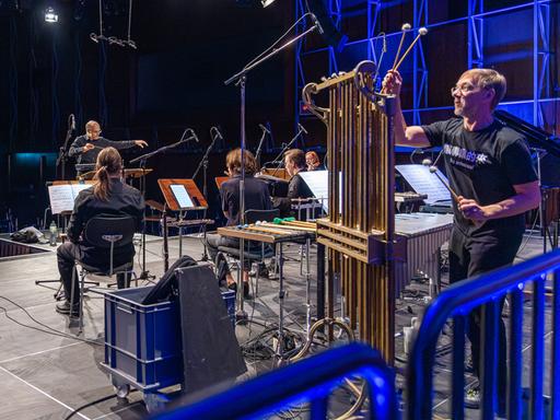 Die Musiker vom E-MEX Ensemble sind zum Teil von hinten, teils von der Seite zu sehen, wie sie gemeinsam spielen. Im Vordergrund stehen große Röhren des Percussions-Musikers.