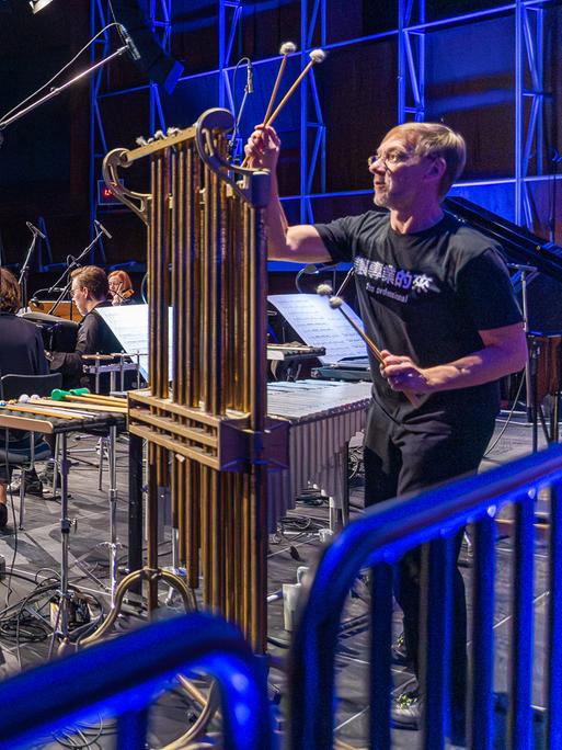 Die Musiker vom E-MEX Ensemble sind zum Teil von hinten, teils von der Seite zu sehen, wie sie gemeinsam spielen. Im Vordergrund stehen große Röhren des Percussions-Musikers.