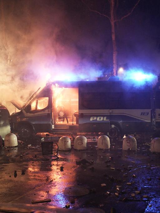 Das Bild zeigt ein brennendes Polizeifahrzeug bei Nacht. Drumherum sind Rauchschwaden und Blaulichter zu sehen.