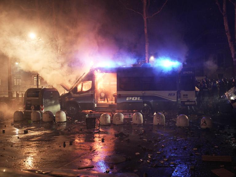 Das Bild zeigt ein brennendes Polizeifahrzeug bei Nacht. Drumherum sind Rauchschwaden und Blaulichter zu sehen.