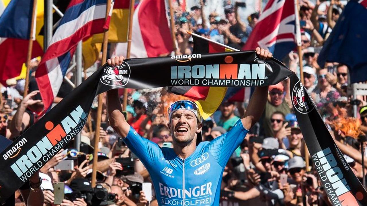 Patrick Lange hält im Ziel ein Ironman-Banner mit beiden Händen über seinen Kopf, im Hintergrund jubelnde Zuschauer und verschiedene Flaggen. Patrick Lange hält im Ziel ein Ironman-Banner mit beiden Händen über seinen Kopf, im Hintergrund jubelnde Zuschauer und verschiedene Flaggen.