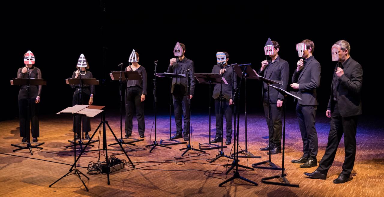 Viele Sängerinnen und Sänger stehen mit Masken vor dem Gesicht im Halbkreis vor einem Dirigentenpult.