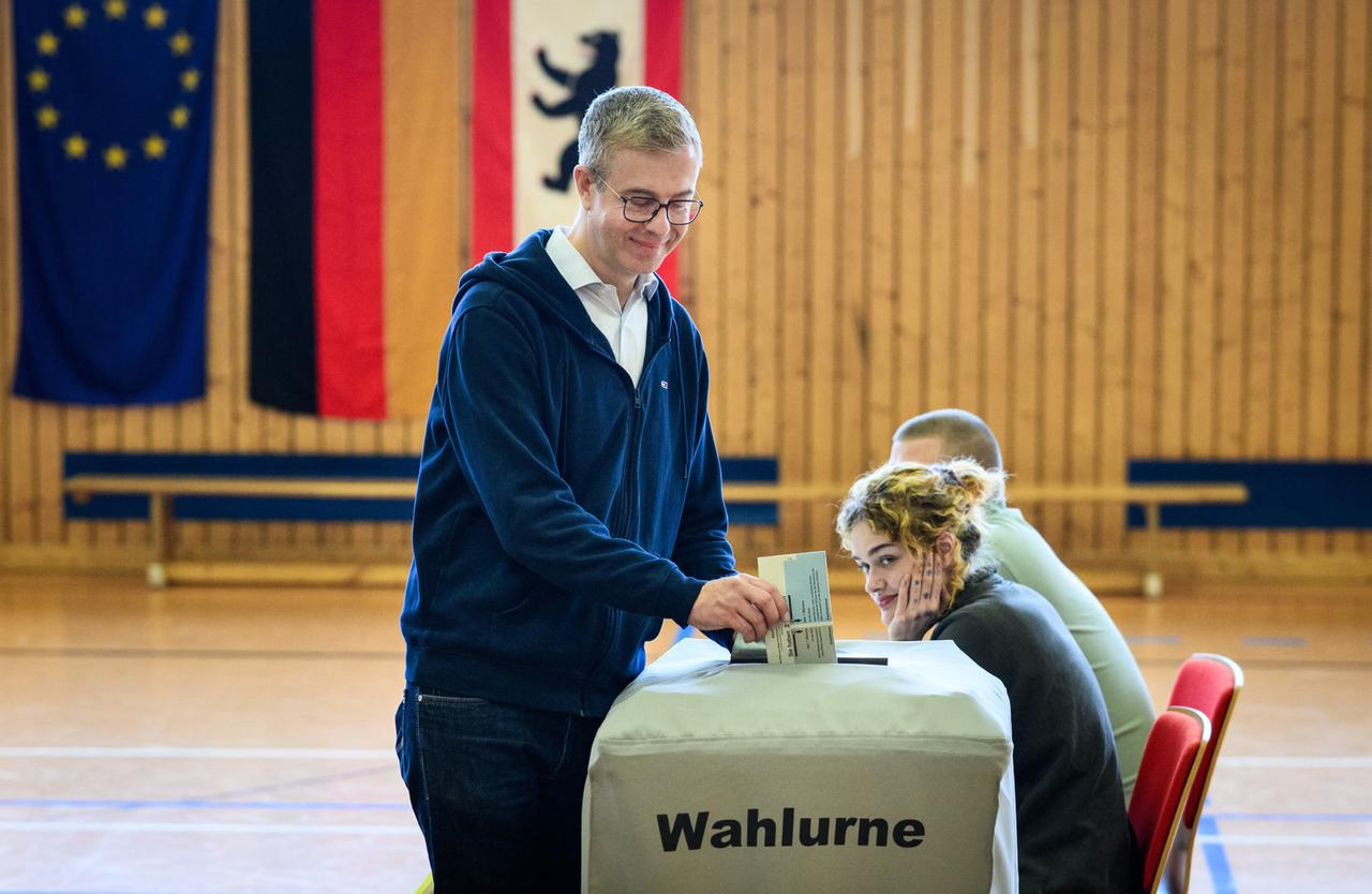 Ein Mann mit Brille, Hemd und Kaputzenpullover wirft einen Stimmzettel in eine Wahlurne.