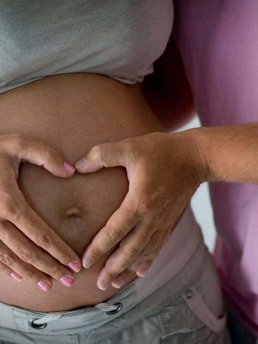 Die Hand einer Frau und die eines Mannes liegen in Herzform auf dem schwangeren Bauch der Frau