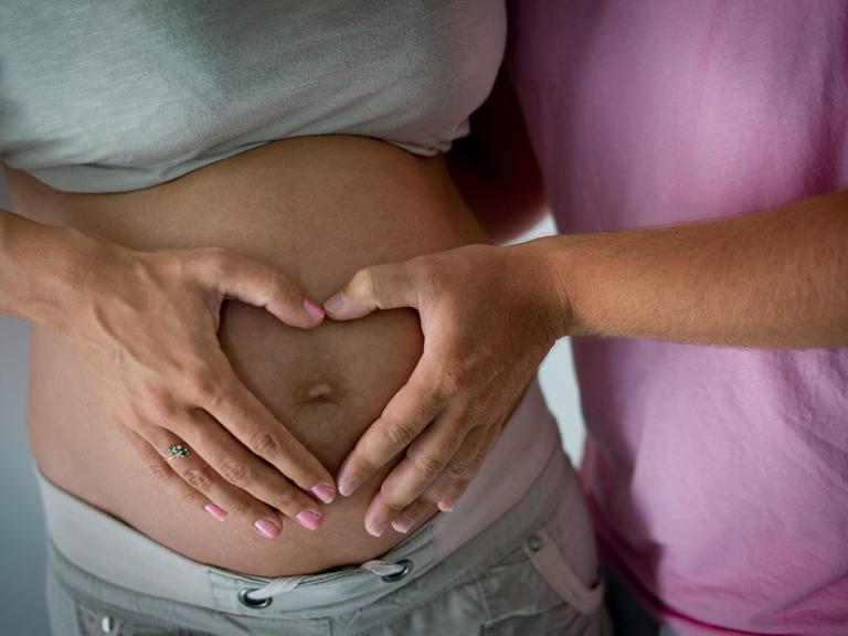 Die Hand einer Frau und die eines Mannes liegen in Herzform auf dem schwangeren Bauch der Frau