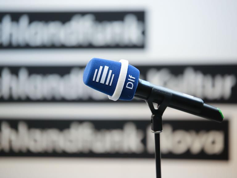 Mikrofon mit Deutschlandfunk-Windschutz vor einer Wand mit den Logos der Sender Deutschlandfunk, Deutschlandfunk Kultur und Deutschlandfunk Nova