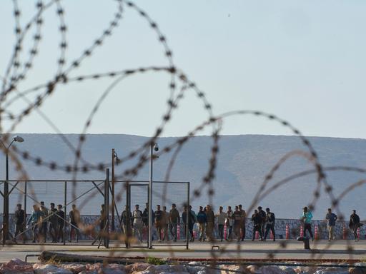 Blick durch einen Stacheldrahtzaun auf eine Gruppe Geflüchtete bei ihrer Ankunft in Griechenland