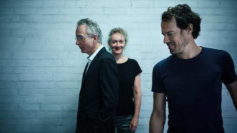 Zwei Männer und eine Frau in ihrer Mitte vor einer weiß gekachelten Wand. 