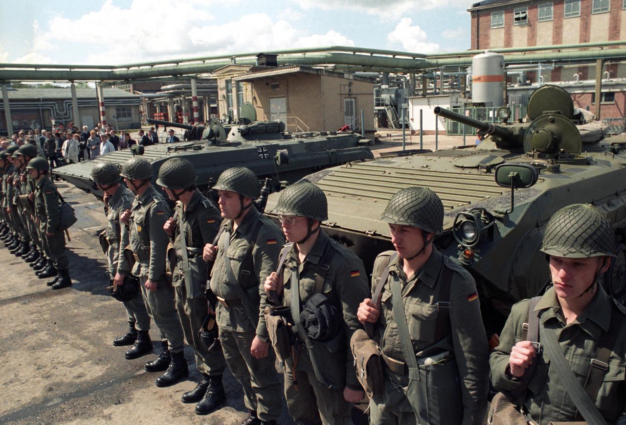 Soldaten stehen vor einem Industriegebäude, vor dem drei NVA-Schützenpanzer stehen.