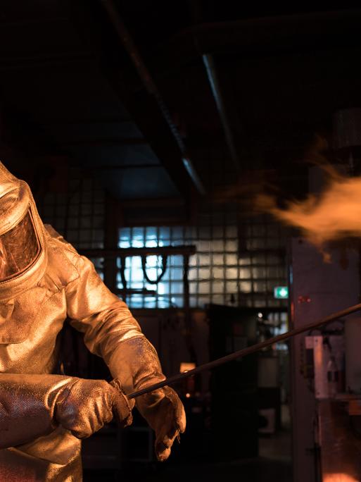 Ein Mitarbeiter der Firma Schott arbeitet an einem Ofen, in dem Glas geschmolzen wird. Er trägt einen Schutzanzug am ganzen Körper, aus dem Ofen schlagen Flammen.