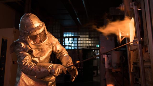 Ein Mitarbeiter der Firma Schott arbeitet an einem Ofen, in dem Glas geschmolzen wird. Er trägt einen Schutzanzug am ganzen Körper, aus dem Ofen schlagen Flammen.