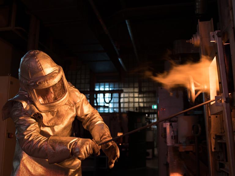 Ein Mitarbeiter der Firma Schott arbeitet an einem Ofen, in dem Glas geschmolzen wird. Er trägt einen Schutzanzug am ganzen Körper, aus dem Ofen schlagen Flammen.