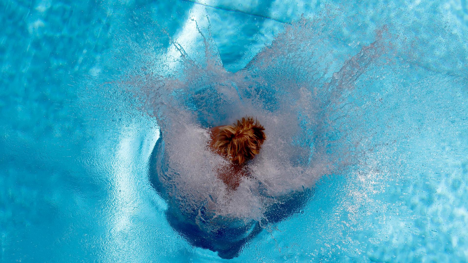 Vogelperspektive auf eine Person, die im Freibad eine Arschbombe ins Becken macht. Wasser spritzt in alle Richtungen.