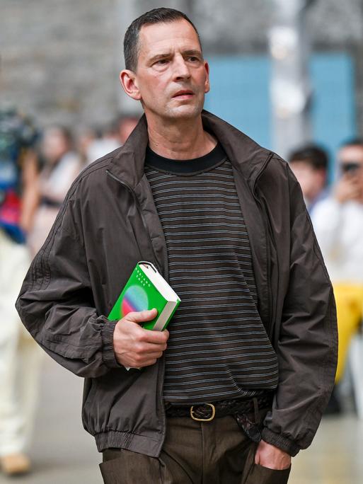 Dunkel gekleideter Mann mit kurzen Haaren und einem Buch unter dem Arm Dunkel gekleideter Mann mit kurzen Haaren und einem Buch unter dem Arm